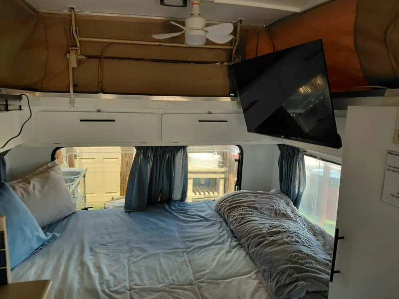 The interior of the caravan showing a made-up double bed under curtained windows with a wall-mounted television