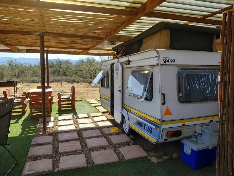 The caravan seen from the side under a shade structure, with a paved path, cooler box and lawn in front of it