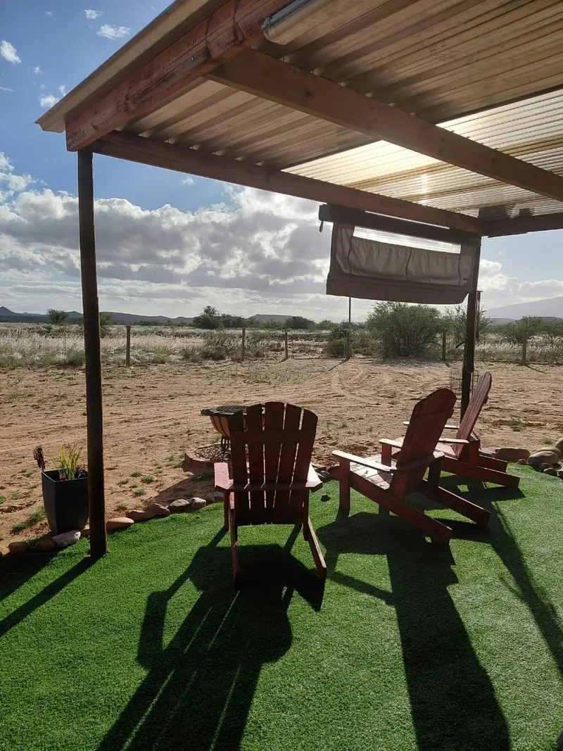 Two red slatted lounge chairs facing out from under a timber pergola towards open veld and blue sky