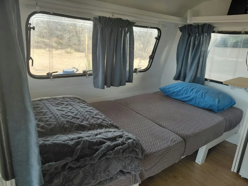 A double bed with a grey blanket and blue cushion inside the caravan, daylight through the side windows