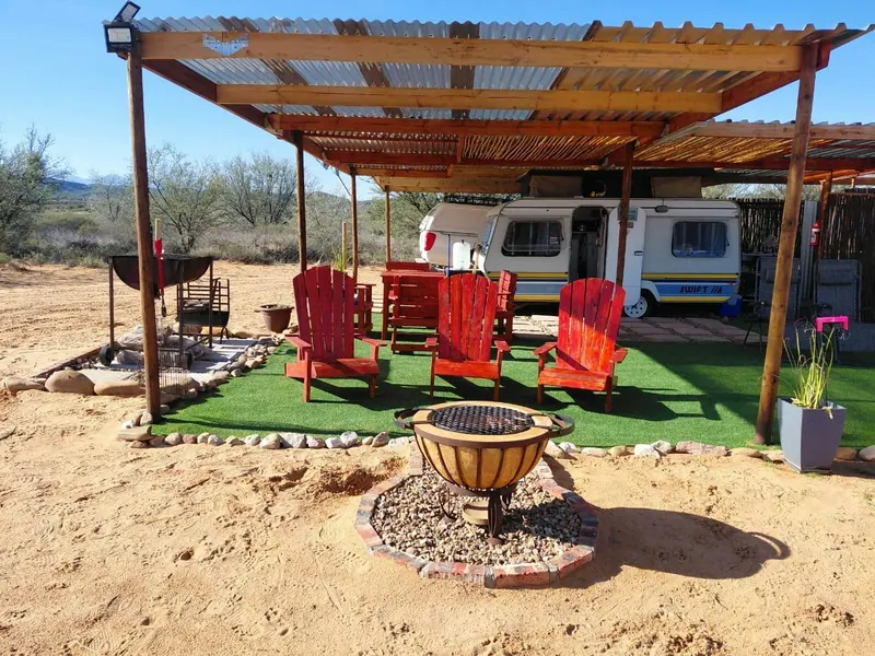 Red timber chairs and a firepit on a lawn under a pergola, with the caravan behind and hills in the distance