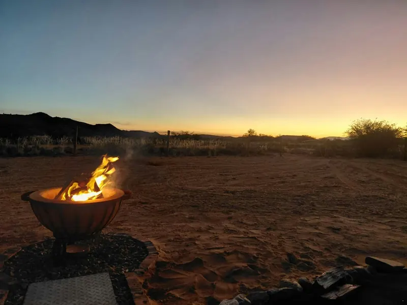 A firepit burning on open red ground at Karoo Stone Camper as the sun sets behind low hills near Ladismith