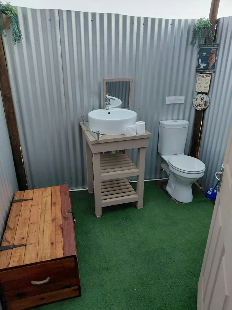 An outdoor bathroom enclosure with a white basin on a timber stand, a mirror and a flush toilet