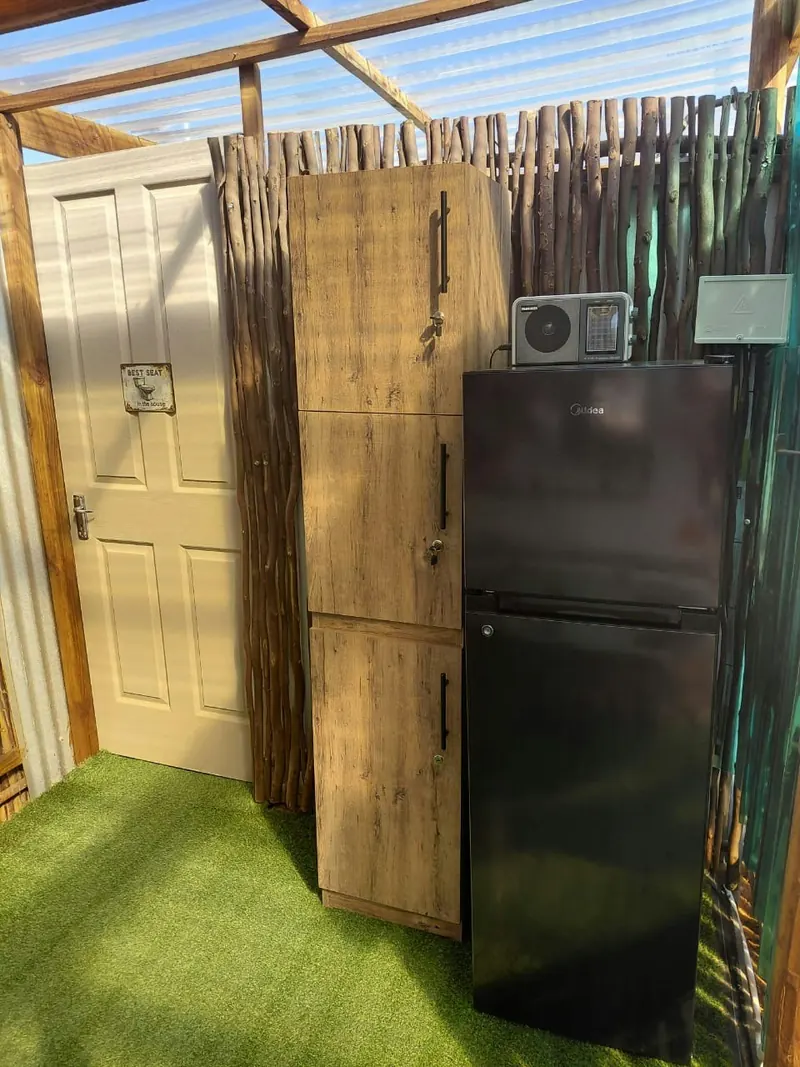 The timber door to the outdoor bathroom standing next to a tall fridge against a reed screen