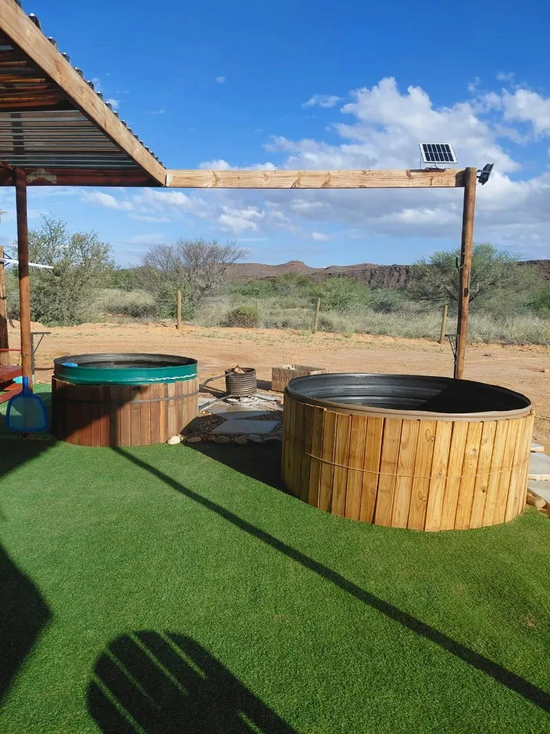 Two timber hot tubs under an open shade frame with a firepit alongside on sandy ground