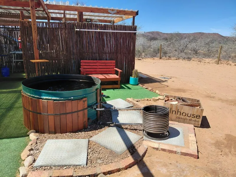 A timber-clad wood-fired hot tub beside a bench and a coiled water heater, looking out over open Karoo veld